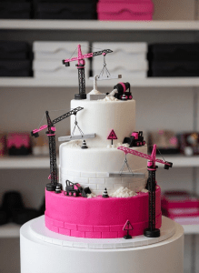 A towering multi-tier custom cake styled like a whimsical construction site, each tier frosted in smooth, pastel buttercream resembling layered building materials. Tiny fondant cranes, bulldozers, and traffic cones perch on ledges, lifting sugar beams and pouring edible “cement.” The cake stands on a clean white pedestal against a softly blurred bakery backdrop with shelves of colorful cake boxes. Gentle studio lighting from the left creates glossy highlights on the icing and subtle shadows under the fondant details. Photographic realism, vibrant and playful, shot at eye level with a shallow depth of field so the construction details are crisp while the background melts into creamy bokeh, capturing the fun “if you can imagine it, we can construct it” spirit.